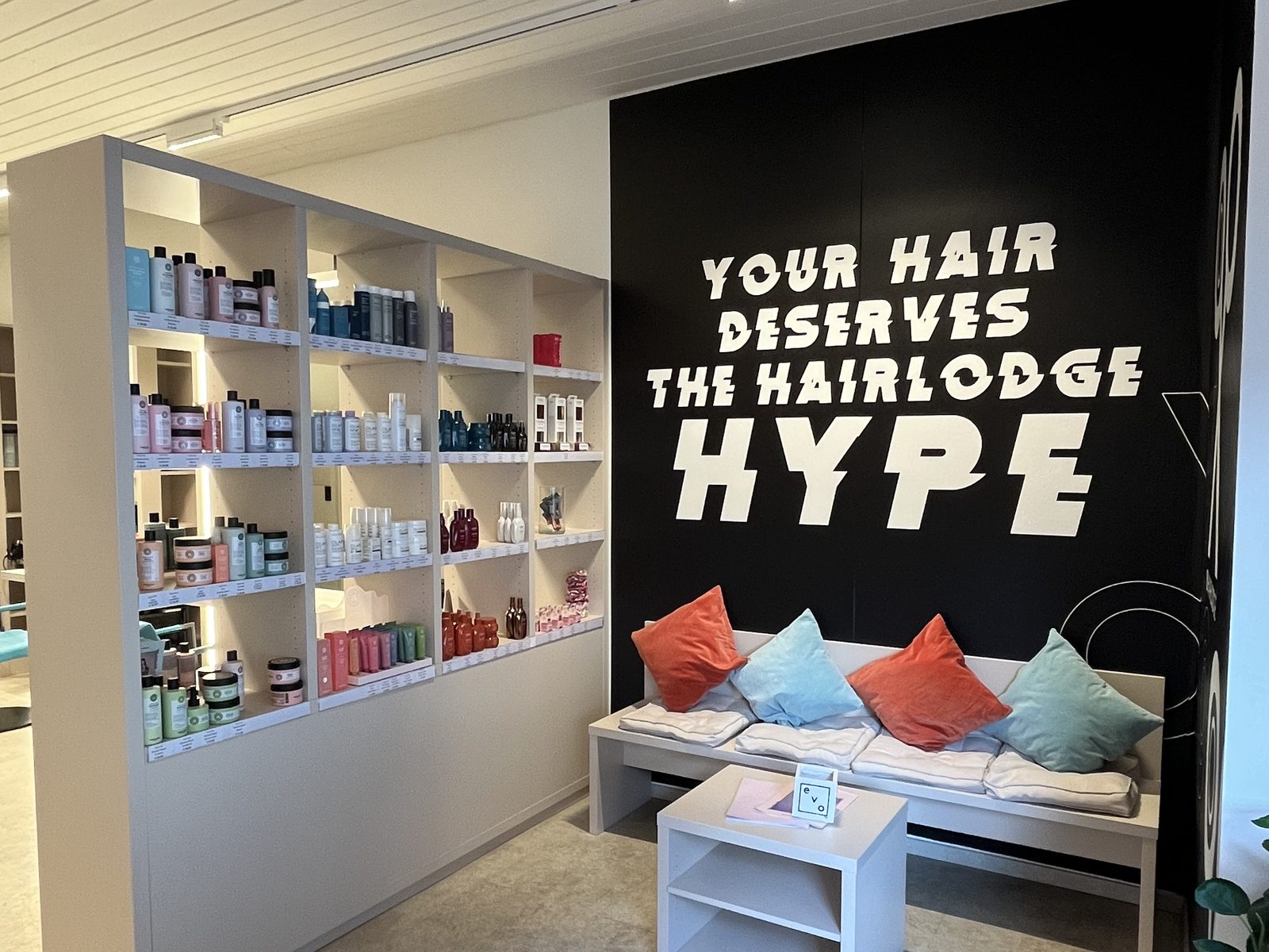 Wartebereich im HAIRLODGE Ellmau: helle Sitzbank mit bunten Kissen vor der schwarzen Wand mit dem Schriftzug "Your hair deserves the Hairlodge Hype", daneben ein Regal voller Pflegeprodukte.