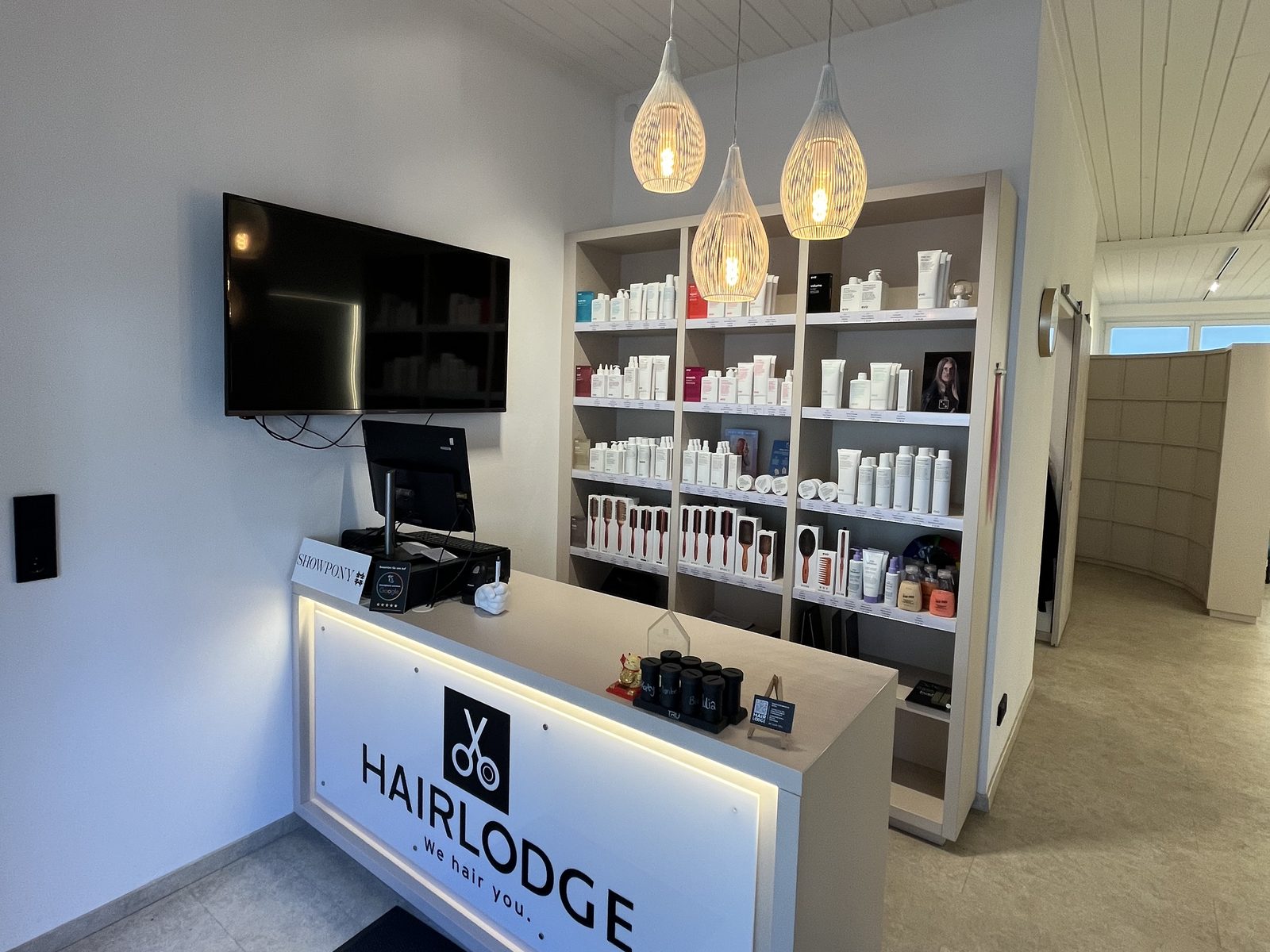 Empfangstresen mit beleuchtetem HAIRLODGE-Logo im Salon Ellmau, darüber drei Pendelleuchten und ein Regal mit Pflege- und Stylingprodukten.