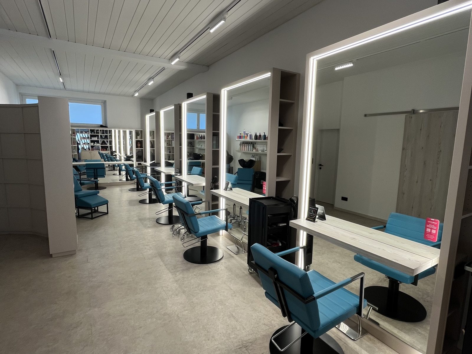 Blick durch den Salon HAIRLODGE Ellmau: petrolfarbene Stylingstühle reihen sich vor den beleuchteten Spiegeln entlang der hellen Wand.