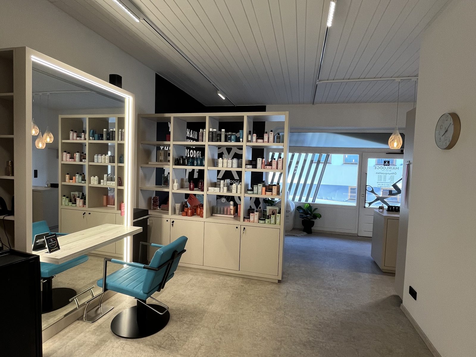 Weiter Blick durch den HAIRLODGE Ellmau bis zum Eingang, mit Produktregal, Stylingplatz und Wanduhr.