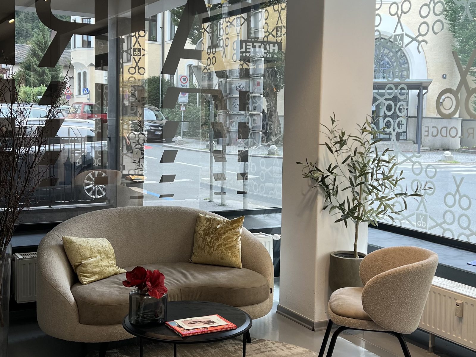Wartebereich im HAIRLODGE Kufstein mit geschwungenem Sofa, Sessel und Olivenbaum vor der bodentiefen Fensterfront.