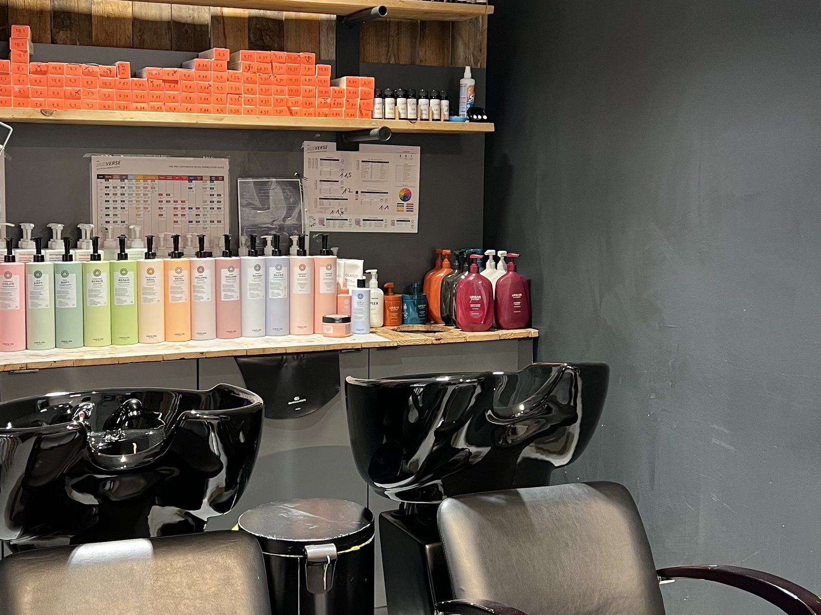 Waschplätze im HAIRLODGE Kufstein vor der anthrazitfarbenen Wand, darüber Farbtuben und Pflegeprodukte im Regal.